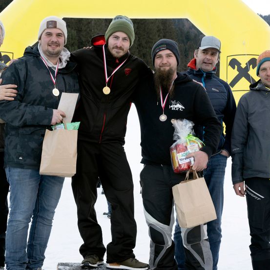 Fünf Männer stehen in einem verschneiten Bereich, drei von ihnen tragen Medaillen. Sie halten Papiertüten, eine davon enthält ein rotes und weißes Paket. Hinter ihnen steht eine gelbe aufblasbare Struktur mit dem Text X.