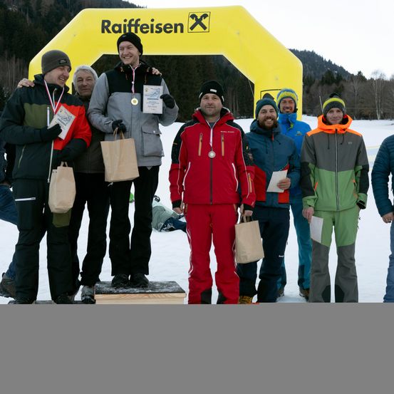 Sieben Männer stehen auf einem verschneiten Feld und halten Medaillen und Papiertüten in den Händen. Sie scheinen Gewinner eines Wettbewerbs zu sein. Sie sind von einem gelben Bogen mit dem Wort Raiffeisen umgeben.