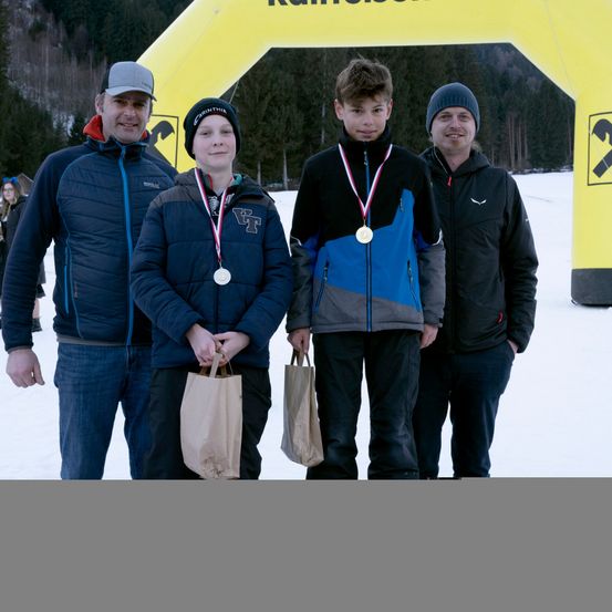 Vier Personen stehen im Schnee und halten Taschen. Zwei Jungen tragen Medaillen. Hinter ihnen steht ein gelber Bogen im Schnee.