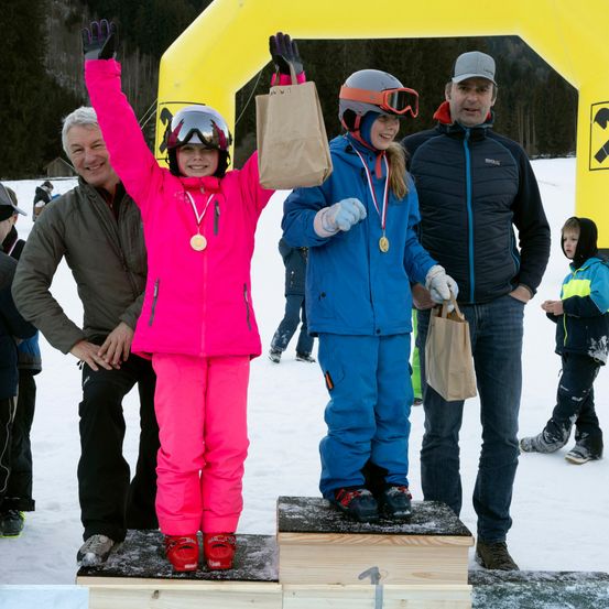 Zwei junge Mädchen in Winterkleidung stehen auf Podests und lächeln, während sie Taschen halten. Sie tragen Medaillen um den Hals. Hinter ihnen halten zwei Erwachsene, einer mit einer grauen Kappe, braune Taschen. Sie befinden sich in einem verschneiten Außenbereich.