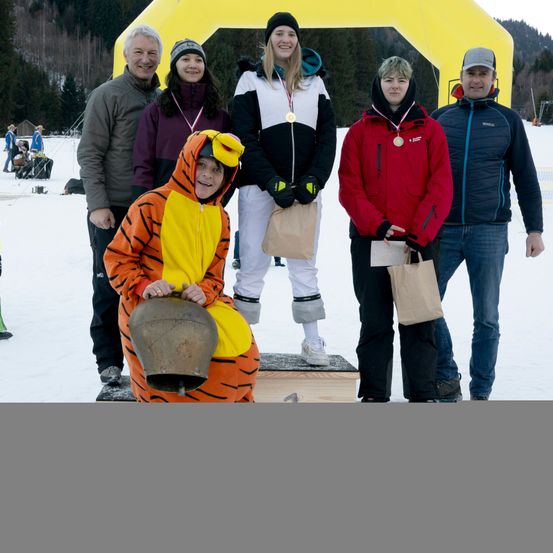 Eine Gruppe von Menschen steht im Schnee. Sie tragen Medaillen und halten Taschen. Eine Person in einem Tigerkostüm hält eine Glocke. Dahinter steht eine gelbe Struktur.