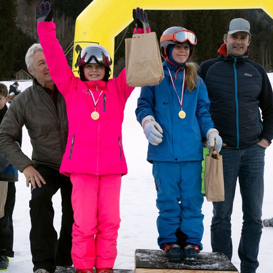 Zwei junge Mädchen stehen auf einem verschneiten Podest, jede hält einen Papierbeutel. Sie tragen beide Medaillen, und zwei Erwachsene stehen hinter ihnen. Der Hintergrund zeigt einen gelben Bogen und verschneite Bäume.