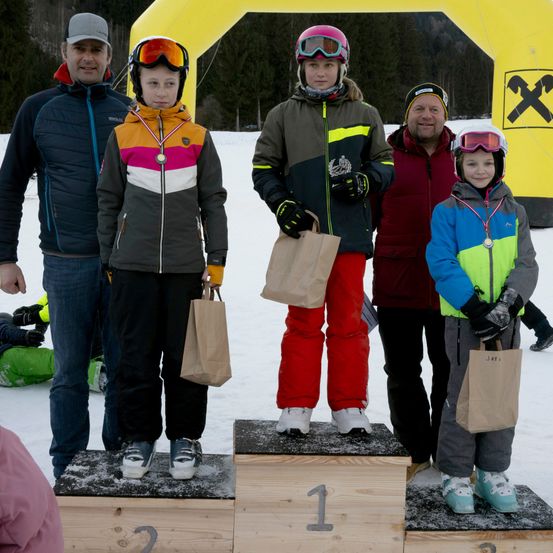 Eine Gruppe von Menschen, darunter junge Kinder, steht auf einem Podest mit den Nummern 1, 2 und 3. Sie sind für Wintersport gekleidet und haben Medaillen um den Hals. Hinter ihnen steht ein gelber Bogen mit einem Hammerlogo.