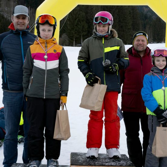 Eine Gruppe junger Skifahrer und ihre Eltern stehen auf einem Podest, halten Papiertüten in der Hand, mit Schnee und einem gelben Bogen im Hintergrund.