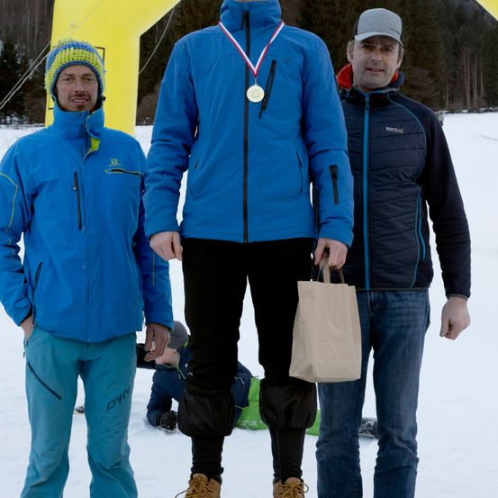 Drei Männer stehen im Schnee, einer trägt eine Medaille und hält einen Beutel. Hinter ihnen steht eine gelbe Struktur und ein verschneiter Hintergrund.