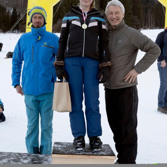 Eine junge Frau steht auf einem Podest in einem verschneiten Bereich, trägt eine Medaille und hält eine Tasche. Sie wird von zwei Männern flankiert, einem in einem blauen und dem anderen in einem braunen Mantel. Bäume und ein gelber Banner sind im Hintergrund.