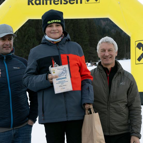 Drei Personen stehen in einem verschneiten Bereich, eine hält ein Zertifikat und einen Papierbeutel, eine andere lächelt, mit Raiffeisen-Banner im Hintergrund.