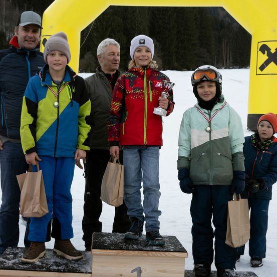 Eine Gruppe von Kindern und Erwachsenen steht auf einem verschneiten Podest mit einem gelben Bogen dahinter. Sie halten Papiertüten und tragen Winterkleidung. Ein Kind hält eine Trophäe.