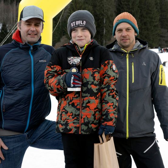 Drei Männer stehen im Schnee. Einer hält eine Trophäe und trägt eine Tarnjacke, während die anderen Mützen und Jacken tragen. Dahinter sind Bäume und ein gelber Bogen zu sehen.