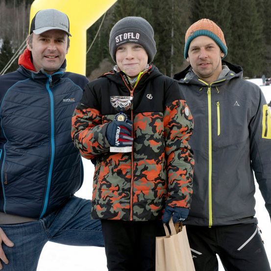 Drei Männer in Winterkleidung posieren für ein Foto. Der Mann in der Mitte hält eine Trophäe, während der Mann auf der rechten Seite einen braunen Papierbeutel hält.