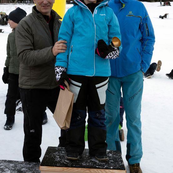 Drei Personen stehen im Schnee, eine auf einem Podest. Die Person auf dem Podest hält einen Becher und trägt Handschuhe. Der Person neben ihm trägt eine blaue Jacke und Hose. Die dritte Person trägt eine schwarze Mütze.
