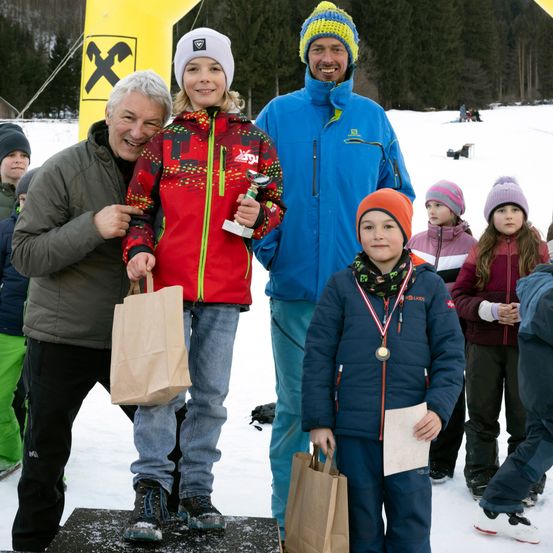 Mehrere Kinder, darunter ein Mädchen mit einem Pokal und ein Junge mit einer Medaille, stehen auf einem Podest mit Erwachsenen. Sie sind auf einem verschneiten Feld mit Bäumen im Hintergrund.