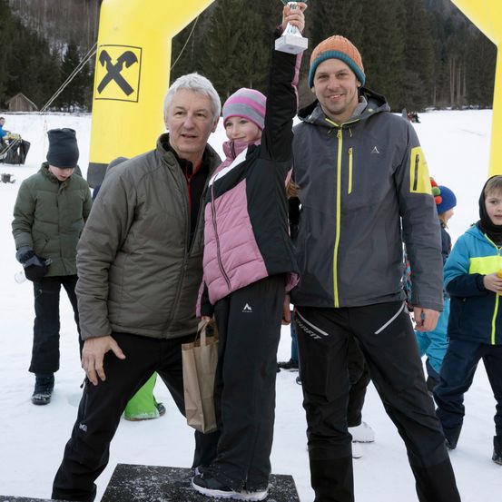 Ein Mann und ein junges Mädchen stehen auf einer verschneiten Plattform und halten eine Trophäe. Hinter ihnen stehen zwei Männer, einer in einem grauen und der andere in einem rosa Jacken. Sie alle lächeln. Dahinter stehen mehrere Leute, einer hält einen Becher. Der Hintergrund zeigt Bäume und einen gelben Bogen.