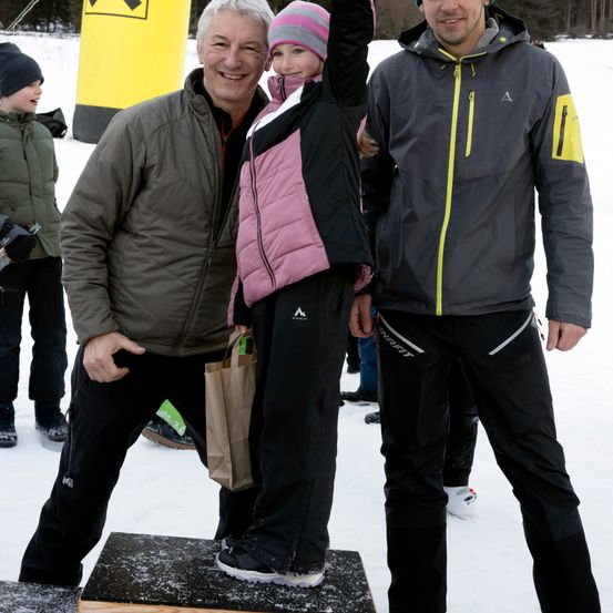 Ein Mann und ein junges Mädchen stehen auf einer Holzplattform im Schnee und halten einen braunen Papierbeutel. Ein anderer Mann steht neben ihnen und lächelt. Sie sind alle für kaltes Wetter gekleidet.