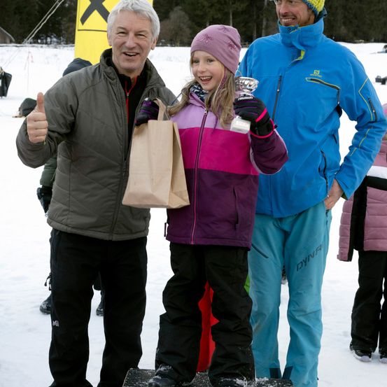 Ein Mann und ein junges Mädchen mit einer Trophäe stehen auf einer verschneiten Oberfläche, mit einem Mann neben ihnen. Dahinter befindet sich eine verschneite Fläche mit Bäumen und einem gelben Schild.