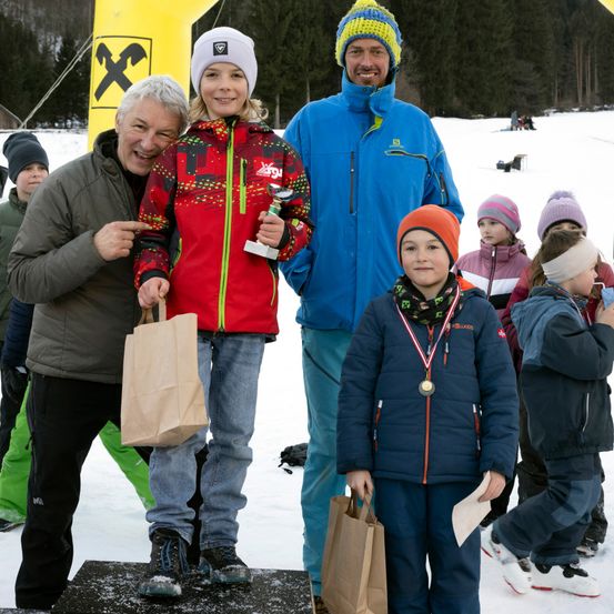Eine Gruppe von Menschen steht auf einer verschneiten Oberfläche. Ein Mädchen mit einer Trophäe und ein Junge mit Medaillen stehen auf einer Plattform. Die beiden Männer neben ihnen halten Papiertüten. Ein gelbes Objekt mit einem Logo ist hinter ihnen.