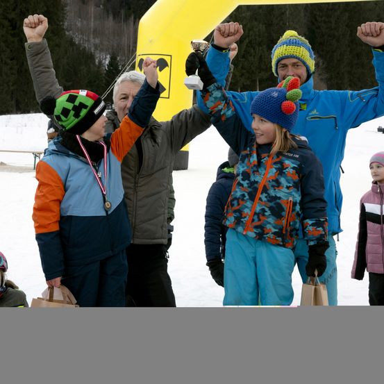 Eine Gruppe von Kindern und Erwachsenen feiert an einem verschneiten Tag, hält Pokale und hebt die Hände. Ein gelber aufblasbarer Bogen ist im Hintergrund.