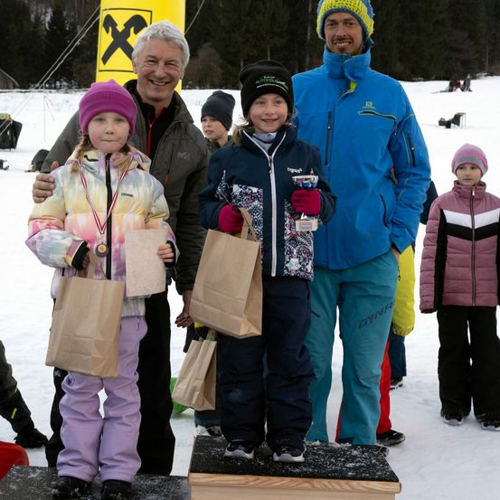 Zwei junge Mädchen stehen auf einer verschneiten Plattform und halten Papiertüten in den Händen, während zwei Männer, einer mit einer Medaille und der andere mit einer Trophäe, hinter ihnen stehen.