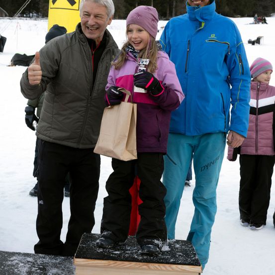 Ein Mann und eine Frau stehen hinter einem jungen Mädchen auf einer verschneiten Plattform, alle lächeln. Das Mädchen hält einen braunen Papierbeutel. Hinter ihnen steht ein weiteres Kind. Bäume und ein gelbes Schild sind im Hintergrund zu sehen.