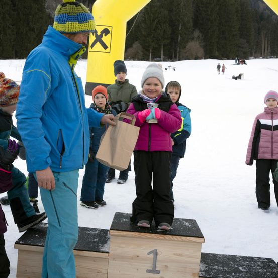 Ein kleines Mädchen mit rosa Handschuhen steht auf einem Podium im Schnee und hält einen Pokal. Ein erwachsener Mann in einem blauen Mantel reicht ihr einen braunen Papierbeutel. Hinter ihnen stehen Kinder und schauen zu.
