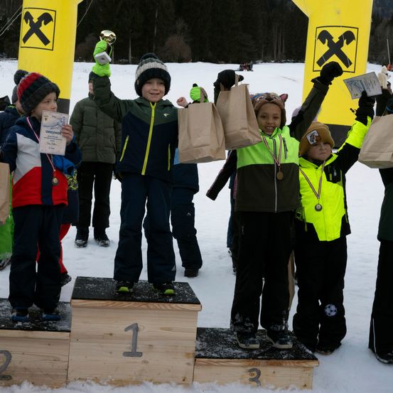 Fünf Kinder stehen auf einem verschneiten Podest, jedes hält einen Papierbeutel und eine Medaille. Der Junge in der Mitte hält einen Pokal und trägt eine Mütze. Dahinter stehen zwei gelbe Aufblasbare mit einem X darauf.
