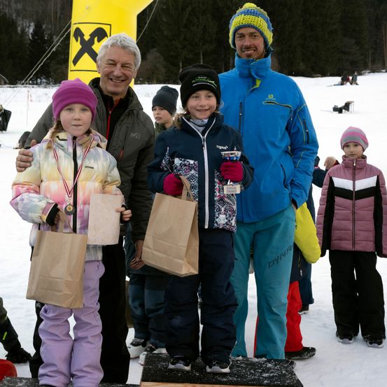 Zwei junge Mädchen stehen auf einem verschneiten Podest mit zwei Männern. Sie halten Papiertüten und Medaillen. Hinter ihnen stehen weitere Kinder im Schnee mit Bäumen und einem gelben Aufblasbaren im Hintergrund.