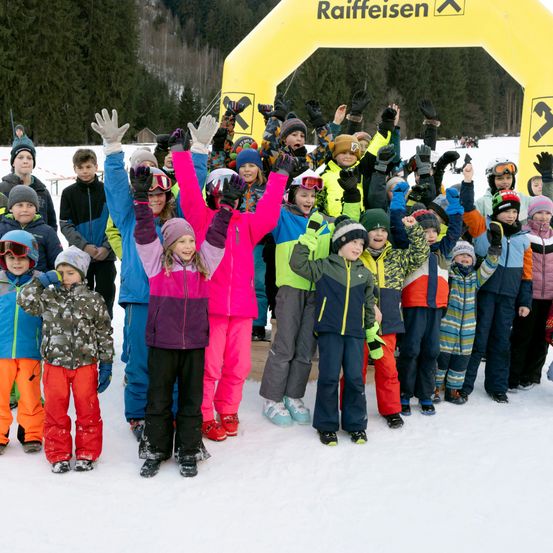 Eine Gruppe von Kindern posiert für ein Foto im Schnee. Sie tragen alle Winterkleidung, einige mit Brillen. Dahinter steht ein gelber Bogen mit dem Wort 'Raiffeisen'.