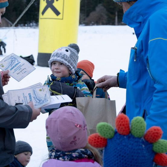 Ein Mann gibt Kindern in Winterkleidung in einer schneebedeckten Außenumgebung Urkunden aus. Die Kinder halten Taschen.
