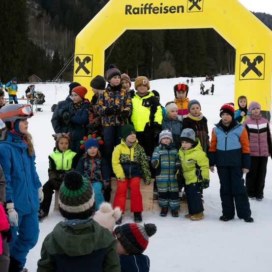 Eine Gruppe von Kindern und Erwachsenen in Winterkleidung steht im Schnee, einige tragen Schutzbrillen und Helme. Sie versammeln sich unter einem gelben aufblasbaren Bogen mit dem Wort Raiffeisen. Hinter ihnen sind Bäume und eine entfernte verschneite Anhöhe zu sehen.