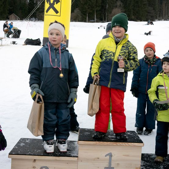 Zwei junge Jungen stehen auf einem Podest, beide tragen Medaillen und halten Papiertüten. Sie sind von anderen Kindern in Winterkleidung umgeben. Dahinter ist ein gelber Banner mit einem schwarzen X zu sehen.