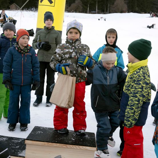 Mehrere Kinder stehen auf einer verschneiten Oberfläche, eines hält einen Papierbeutel. Sie tragen Winterkleidung, und ein gelber Schlauch ist hinter ihnen.