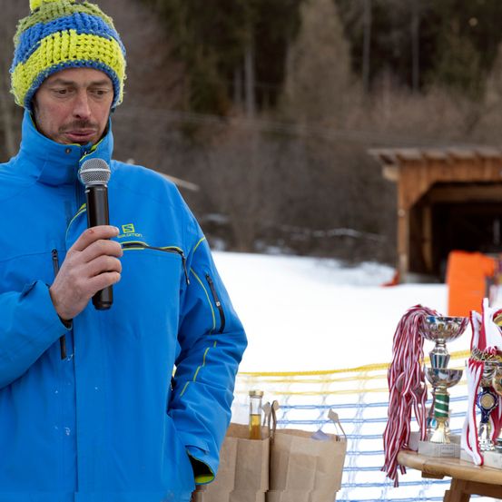 Ein Mann in einem blauen Mantel und einer bunten Mütze spricht in ein Mikrofon. Er steht im Schnee, mit Trophäen und einer Holzstruktur im Hintergrund.