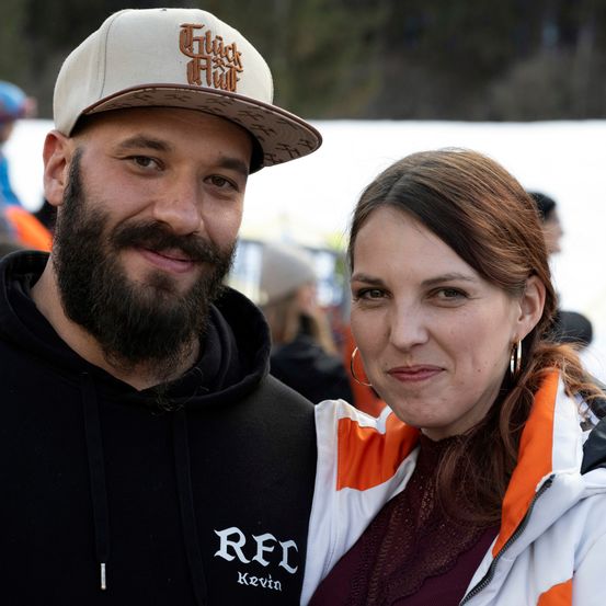 Ein Mann mit Bart und eine Frau stehen nah beieinander und lächeln für die Kamera. Der Mann trägt eine Kappe und eine schwarze Kapuzenjacke, während die Frau eine weiße und orangefarbene Jacke trägt. Sie scheinen bei einer schneebedeckten Veranstaltung zu sein.