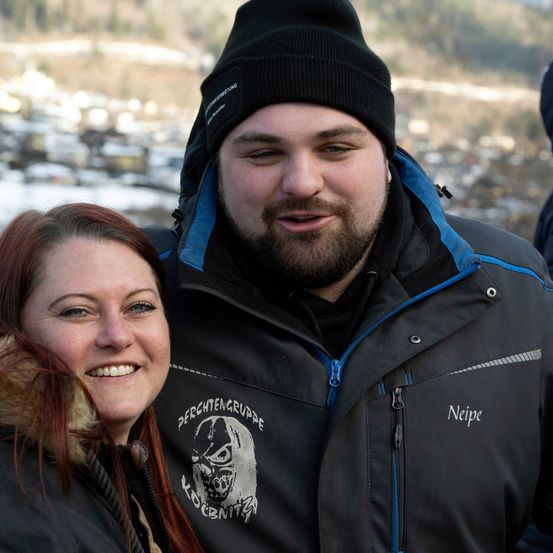 Ein lächelndes Paar steht eng zusammen, mit einem verschneiten Hintergrund. Der Mann trägt eine schwarze Mütze und eine Jacke mit dem Wort Neipe.