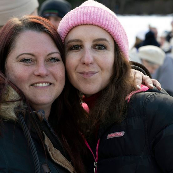 Zwei Frauen lächeln in die Kamera, eine mit langen Haaren und einer rosa Mütze, die andere mit einer pelzgefütterten Jacke und dunklen Haaren. Sie stehen eng beieinander und haben ihre Arme umeinander geschlungen.