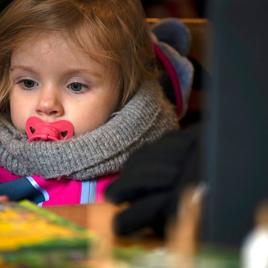 Ein kleines Mädchen mit blonden Haaren, das einen rosa Schnuller und einen grauen Schal trägt und geradeaus schaut. Sie trägt eine rosa Jacke mit blauen Streifen.
