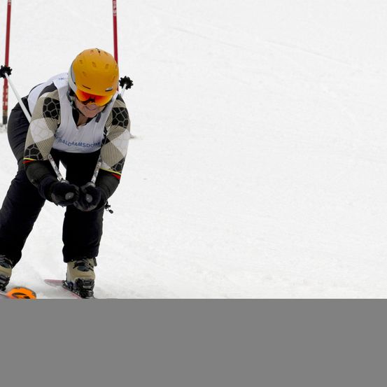 Ein Skifahrer mit gelbem Helm und Brille fährt den Schneehang hinunter und hält Skistöcke.