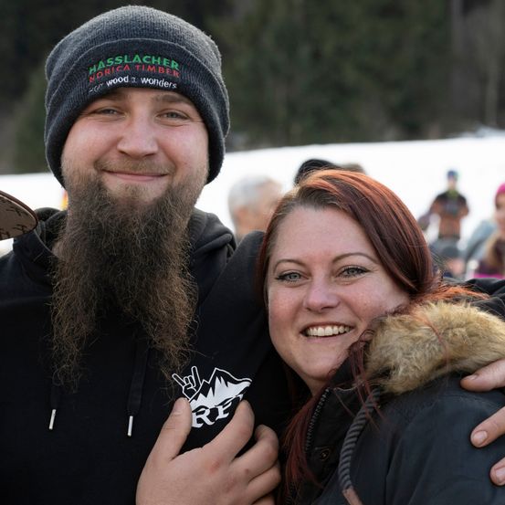 Ein lächelnder Mann und eine Frau, beide in Winterkleidung, stehen eng beieinander mit dem verschneiten Hintergrund.