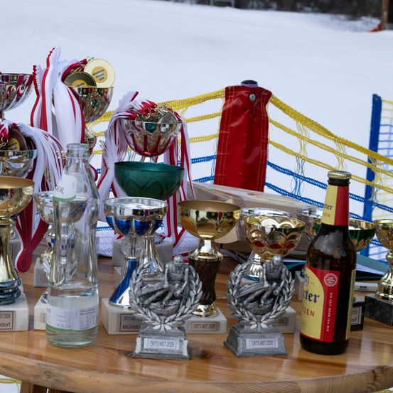 Verschiedene Trophäen und Medaillen sind auf einem Holztisch ausgestellt, darunter eine Flasche Bier. Hinter dem Tisch befindet sich ein Netz, und im Hintergrund ist schneidebedeckter Boden zu sehen.