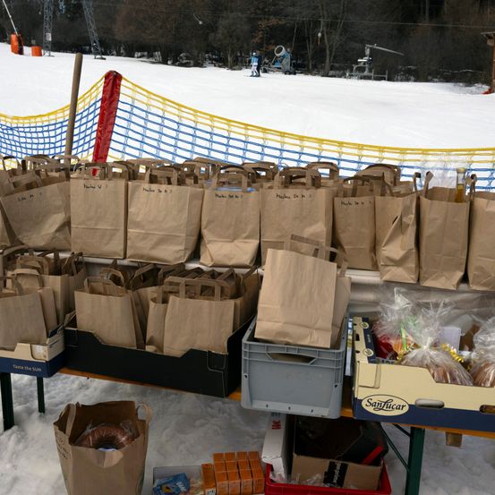 Viele braune Papiertüten stehen auf einem Tisch im Schnee. Ein Kasten mit SanLucar-Brot und anderen Gegenständen steht ebenfalls auf dem Tisch.