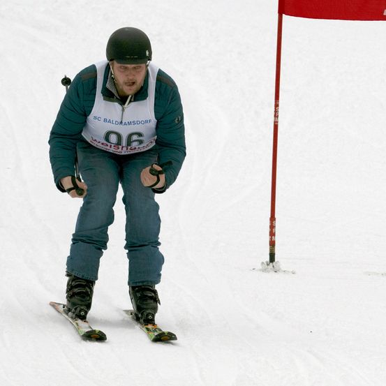 Ein Skifahrer mit Helm und Skischuhen fährt bergab auf einer verschneiten Piste. Er hält Skistöcke und trägt ein Trikot mit der Nummer 96. Ein roter Flaggenpfosten steht im Schnee hinter ihm.