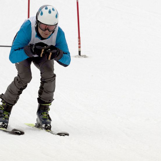 Ein Skifahrer in blauer Jacke und grauer Hose fährt den Schneehang hinunter. Er trägt einen weißen Helm, eine Brille und Handschuhe.
