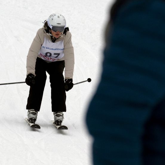 Ein Skifahrer mit weißer Helm und Schutzbrille, Nummer 87, fährt den Schneehang hinunter und hält Skistöcke.
