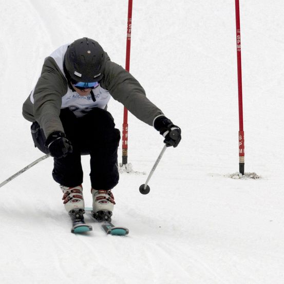 Ein Skifahrer mit Helm und Schutzbrille navigiert durch eine Slalomstrecke und fährt zwischen roten Stangen auf einer verschneiten Piste.