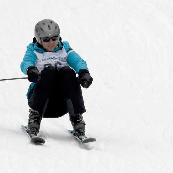 Ein Skifahrer in einer blauen Jacke mit SC Baldramsdorfe darauf skisst einen schneebedeckten Hang hinunter und trägt einen grauen Helm und schwarze Handschuhe.
