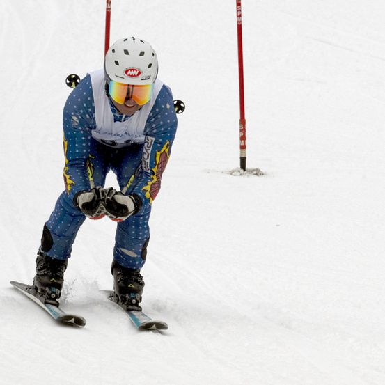 Ein Skifahrer fährt auf einem verschneiten Hang, trägt einen Helm und eine Brille. Er beugt sich leicht nach vorne und hält seine Skistöcke.