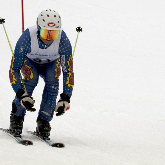 Ein Skifahrer mit weißem Helm und Schutzbrille fährt einen schneebedeckten Hang hinunter und trägt blaue und weiße Skiausrüstung.