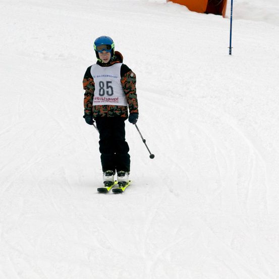 Ein junger Skifahrer mit der Nummer 85 auf der Jacke fährt einen verschneiten Hang hinunter, trägt einen Helm und eine Schutzbrille.
