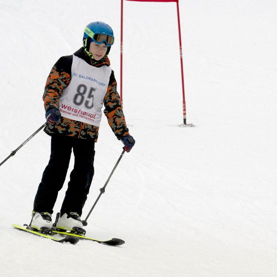 Ein Skifahrer mit einer Camouflage-Jacke und einem Startnummer-85-Startertrikot, der einen verschneiten Hang hinunterfährt, mit roten Pfosten im Hintergrund.