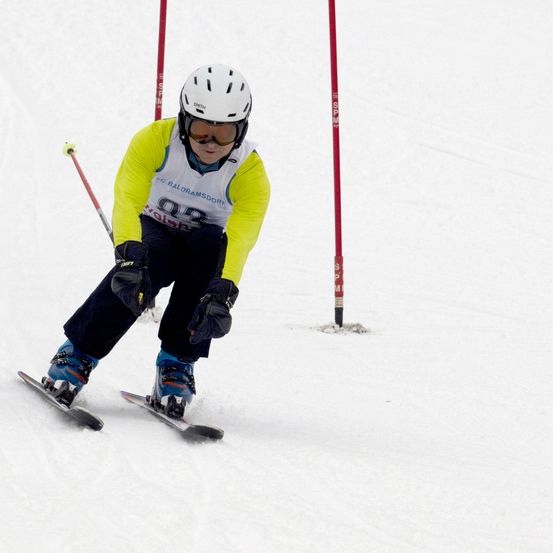 Ein Skifahrer in einem gelben und weißen Skianzug mit der Nummer 93 fährt auf einem verschneiten Hang. Der Skifahrer hält Skistöcke und trägt einen weißen Helm.